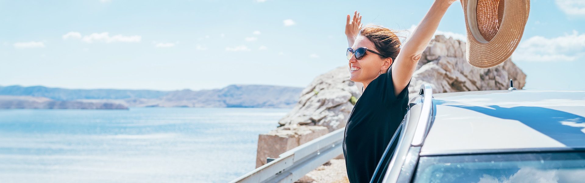 Cheerful Woman portrait enjoying the seaside road trip. Dressed a black dress with straw hat and sunglasses she wide opened arms and shining with happiness. Summer vacation traveling by auto concept.