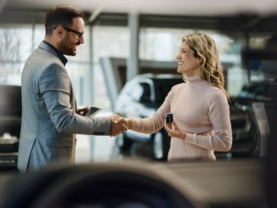 Happy salesman congratulating his female customer for buying a new car in a showroom. The view is through glass.